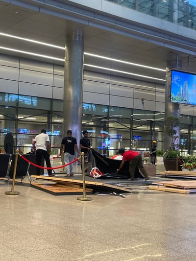 The image shows a group of men setting up a display or event in a modern, indoor public space, possibly an airport or convention center. The architecture features polished floors, large glass windows, and prominent cylindrical columns. Several men are actively engaged in the setup. One man in a red shirt is seen pulling a large black cloth, possibly a backdrop or cover, while another man in a black t-shirt holds a trash bag. Two other men in casual attire are walking away from the setup area, one in a white t-shirt and black pants, and another in a black t-shirt and light-colored pants. In the background, a large digital billboard displays an advertisement for "IMPERIA," featuring images of modern buildings. A few potted plants and red velvet ropes with gold stanchions are also visible, indicating an organized event space. The overall scene suggests preparation or dismantling of an installation. The location is identified as Gollapalle Khurd, India.