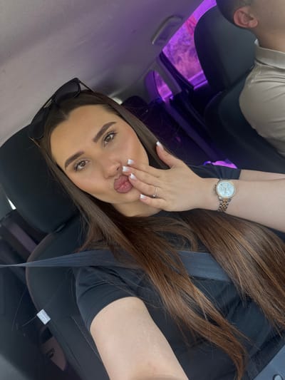 A young woman with long brown hair is in the passenger seat of a car, giving a kiss to the camera. She is wearing a black t-shirt and a watch on her left wrist. Her right hand is held up to her face, and she is wearing a ring on her left ring finger. The interior of the car is visible, with the headrest of the driver's seat and a purple-tinted window in the background. The scene appears to be during daylight hours.