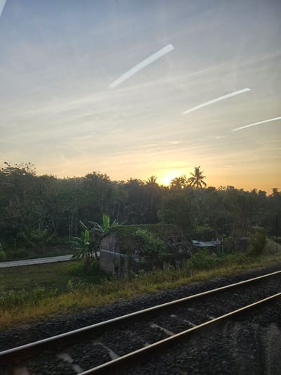 This is a photograph taken from a moving train in Kedungsari, Indonesia, looking out a window. The scene depicts a rural landscape at sunset. The sky is a gradient of soft blues and yellows, with several white, rectangular reflections on the window glass, possibly from interior lighting. Below the sky, a dense line of trees, including palm trees, forms a silhouette against the horizon, with the setting sun visible as a bright orange orb peeking through the foliage. In the mid-ground, there is a dilapidated brick building with a roof covered in greenery. Adjacent to the building is a road and a grassy area with some vegetation. In the foreground, two train tracks run diagonally across the bottom of the frame, blurred due to the motion of the train. The overall impression is one of a tranquil, natural setting at dusk, observed from the perspective of travel.