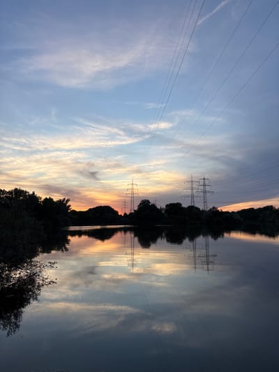 The image depicts a tranquil natural scene at dusk, likely captured in Ludwigshafen am Rhein, Germany, given the presence of pylons and the landscape composition. A body of water occupies the foreground, reflecting the sky and the surrounding environment. The sky is a canvas of soft hues, transitioning from a pale blue at the top to warmer oranges and yellows near the horizon, indicating the setting sun. Wispy clouds are scattered across the sky, adding texture and depth.
Dominating the middle ground are silhouetted trees lining the opposite bank of the water. Emerging from behind the trees are several large electricity pylons, typical of German infrastructure. These structures are starkly outlined against the colorful sky and also mirrored in the calm water. Overhead, power lines stretch across the frame, further emphasizing the man-made elements within the natural setting. The overall atmosphere is one of peaceful twilight, with the natural beauty of the sunset complemented by the industrial presence of the pylons. No people are visible in the image.