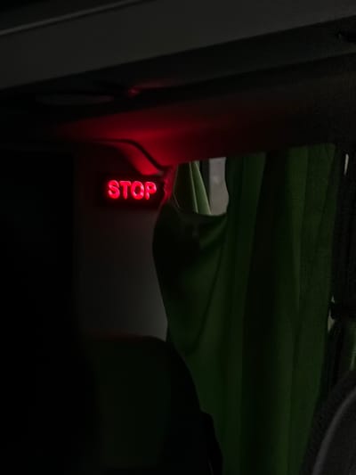 An illuminated red "STOP" sign is visible within the interior of a vehicle located in Moineville, France. The sign, rendered in capital letters, glows brightly against a predominantly dark environment. It is mounted on an overhead panel or compartment. Thin black wiring extends from the top of the sign into the overhead structure. To the right of the sign, a section of a green fabric curtain hangs vertically, partially obscuring a lighter area or window beyond. The foreground and bottom-left portion of the image are obscured by dark, indistinct shapes, consistent with vehicle seating or interior components. No individuals or specific actions are directly observed. The scene is characterized by low ambient light, emphasizing the illuminated "STOP" indicator.