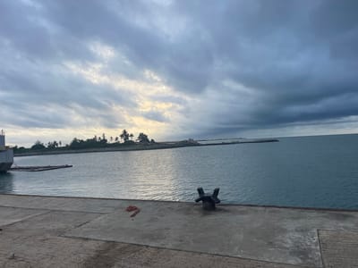 The image shows a concrete pier in Ebute Leki, Nigeria. In the foreground, there is a weathered concrete surface with a metal mooring bollard and a coiled rope. Beyond the pier, there's a calm expanse of water, with a low-lying landmass in the distance covered with trees and vegetation. The sky is heavily overcast with dark, ominous clouds. No people are visible. A ship is partially visible on the left side of the image.
