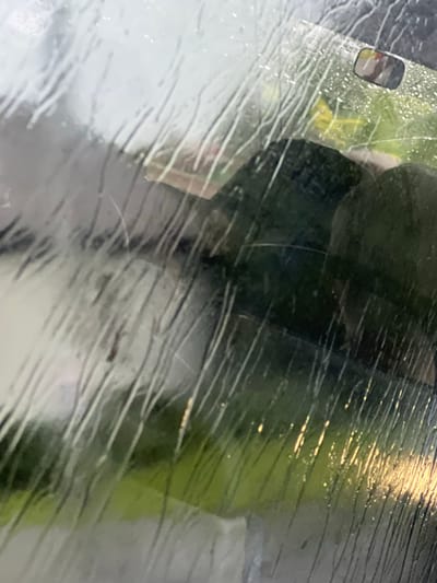 The image is a close-up of a car windshield with raindrops on it, distorting the view of the outside. The raindrops create streaks and a blurred effect, making it difficult to discern specific details of the environment. Through the rain-streaked glass, a reflection of a rearview mirror can be seen, showing a partial view of what appears to be a person's head inside the car. Beyond the windshield, there is a blurred landscape with green and yellow hues, suggesting vegetation or possibly a road with streetlights. The overall impression is of a rainy day in a potentially urban or suburban setting, but no specific landmarks or features are identifiable to pinpoint the exact location within Rzędzianowice, Poland. The lighting appears to be overcast, typical of rainy weather.
