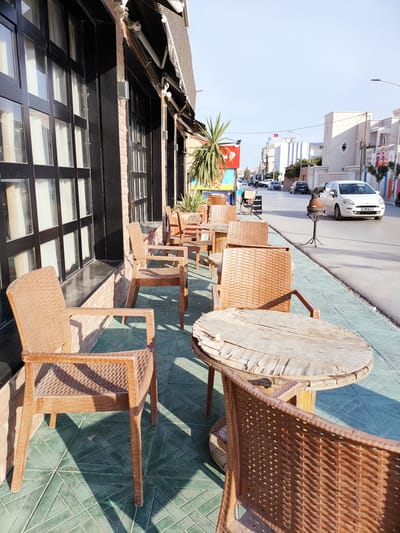 This media file shows an outdoor seating area of a cafe or restaurant in Ariana, Tunisia. The scene is set on a sidewalk adjacent to a building with large, dark-framed windows. Several tables and chairs are visible, predominantly made of brown wicker or rattan-like material, with one rustic wooden table in the foreground. The chairs have a woven pattern and armrests.
The setting appears to be a street with moderate traffic, including several cars driving along the road. Buildings line the opposite side of the street, some of which are white with distinctive architectural features. A Tunisian flag is visible on a building in the distance. The ground is paved with green tiles forming a geometric pattern, and shadows from the tables and chairs are cast upon it, indicating it is daytime.
The time of day appears to be late morning or early afternoon, judging by the angle and length of the shadows and the clear, bright sky. The weather seems pleasant, with sunshine and a clear sky. The overall mood is calm and ordinary, suggesting a typical day for a street-side establishment. There is some visible text on a sign in the background, but it is illegible in this image. A palm-like plant is also present, adding to the Mediterranean feel of the location. The scene captures a moment of everyday life in a Tunisian urban environment.