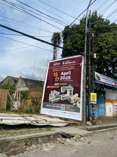 A large billboard with a red background stands on a sidewalk in the city of Kecamatan Dayeuhkolot, Indonesia. The billboard advertises the "Indonesia International Textile & Garment Industry Exhibition" known as "Inatex - Indo Intertex" and is scheduled for April 15-17, 2025 at the "Jakarta International Expo". There is a sewing machine and spools of thread pictured next to a piece of white fabric. The billboard lists the phone number "+62 821-2000-9200" and the website "indointertex.com" for further information. There is a QR code to the right of the phone number. The billboard also states "Register Your Free Visit!".
The billboard is mounted on a wooden post with power lines running above. A tree with green leaves is behind the billboard. There is a small wooden fence and a bamboo mat on the ground in front of the billboard. A brick wall and wooden door are to the right of the billboard, along with another sign. The sidewalk is uneven and cracked. The sky is partially obscured by the trees and power lines.