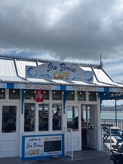 The image depicts the exterior of the "Sea Breeze CAFE" in Llandudno, United Kingdom. The cafe is a white building with blue accents and is adorned with signs advertising hot chocolate, tea, biscuits, and coffee. One sign prominently displays "Sea Breeze CAFE" along with wave motifs. Another sign lists "Daily Specials," including Hummingbird and Cherry Blossom options. Through the windows, people can be seen seated inside. The cafe appears to be situated on a pier or boardwalk, with the sea and the Llandudno coastline visible in the background. The sky is overcast with heavy grey clouds.