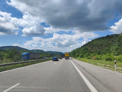 The image depicts a view of a highway in Vrachesh, Bulgaria, on a cloudy day. The road, paved with asphalt, has two lanes in the direction of travel, marked by a solid white line on the right and a broken white line on the left. A guardrail runs along the left side of the road, separating it from the adjacent embankment.
On the highway, there are several vehicles. In the foreground, on the left lane, a blue sedan is traveling. Further ahead, in the right lane, a yellow truck with a trailer is visible, and behind it, another vehicle. On the left side of the highway, a blue semi-trailer truck is parked or moving slowly on what appears to be an access road or a service area.
The environment surrounding the highway is characterized by rolling hills covered in dense green trees. The sky is partially covered with large, white cumulus clouds, interspersed with patches of blue sky. The overall impression is one of a rural highway scene under dynamic weather conditions.