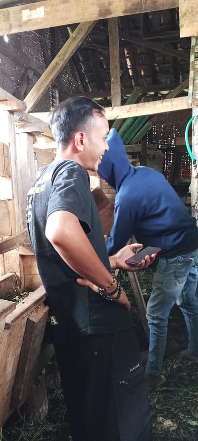 The image shows two men standing in a wooden structure. The man on the left is wearing a black t-shirt with a white logo and black pants. He is smiling and holding a black smartphone with a red case in his right hand. The man on the right is wearing a blue hoodie and blue jeans. He is looking down at the phone in the man on the left's hand. The men are standing in front of a wooden structure that appears to be a barn or shed, with wooden beams and a thatched roof. The ground is covered in green foliage. The location is in the city of Cimaung, Indonesia, based on the coordinates (latitude: -7.086, longitude: 107.576).