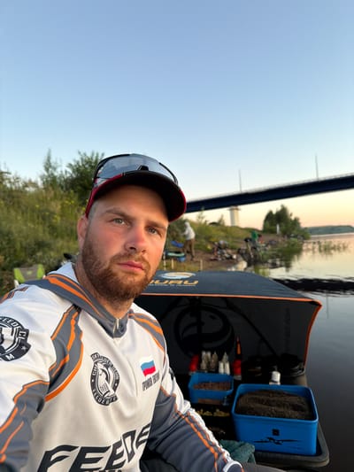 The image shows a man wearing a fishing vest and a baseball cap, with sunglasses perched on the cap. He is looking at the camera. In the background, there is a body of water, a bridge, and trees. The man appears to be at a fishing spot. The location is identified as Tarasovo, Russia. The scene is set in what appears to be a natural environment, possibly a riverbank or lakeside, with the sky suggesting it is either sunrise or sunset. Several other people are visible in the background, also seemingly engaged in fishing activities, with fishing gear and tents set up along the shore. There are containers of fishing bait and other supplies near the man.