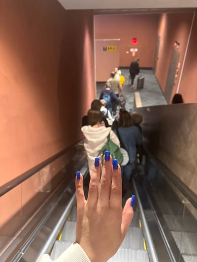 In Yuzhnyye Koryaki, Russia, on an escalator, a person's hand with bright blue fingernails is in the foreground, obscuring the view of the escalator steps. Behind the hand, a group of people are ascending the escalator, each carrying luggage. The environment is enclosed with brown walls, and signs are visible on the wall.