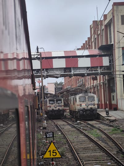 The image captures a scene at the Bhopal Junction railway station in Bhopal, India. In the foreground, the side of a red and white striped train carriage is visible, reflecting the surrounding environment. Two electric locomotives, identified as WAP 7 and bearing the number 37256, are positioned on parallel railway tracks. The locomotives are cream-colored with red and brown accents and feature the Indian flag emblem.

Overhead, a pedestrian bridge with alternating red and white striped awnings spans the tracks, connecting buildings on either side. The buildings themselves are a mix of architectural styles, with one prominently featuring red columns and cream-colored walls, interspersed with windows. Electrical wires and support structures for the overhead power supply for the trains are also visible.

On the ground, numerous railway tracks are present, with ballast and scattered debris. A yellow triangular sign with the number "15" in black is mounted on a pole, indicating a speed limit. Another black and white rectangular sign reads "LWR NO.20". A red "STOP" sign is also visible on a pole. The sky is overcast and grey. The overall atmosphere suggests a busy railway station environment.