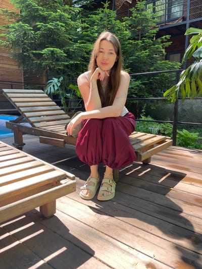 A young woman with long brown hair is sitting on a wooden lounge chair on a wooden deck. She is wearing a light-colored top and loose, dark red pants that gather at the ankles. She has on light-colored sandals and a bracelet on her left ankle. Her right hand is resting on her chin, and she is looking at the camera. Behind her, there are evergreen trees, a wooden building with balconies, and a metal railing. In the foreground, another empty wooden lounge chair is visible. The lighting suggests it is daytime, with shadows cast by the lounge chairs. The scene appears to be an outdoor recreational area, possibly at a hotel or resort, in Guzeripl', Russia.