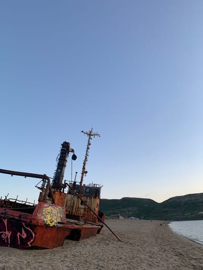 An abandoned, rusted shipwreck is beached on a sandy shore. The ship, adorned with graffiti, lies on its side. In the background, green, treeless hills rise above a calm body of water. A few small figures are visible walking on the beach and near some buildings nestled into the hillside. The sky is clear and a pale blue, indicating it is either early morning or late evening. This scene is located on the coast of Teriberka, Russia.