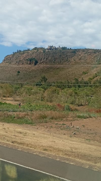 A landscape features a brown hill topped with buildings under a cloudy sky. A lower slope leads to vegetation and what appears to be a garden with someone working. Wires stretch across the middle ground. A dry, grassy area stretches in the foreground and a road with a white line is in the very bottom. The photo is likely taken from a moving vehicle.
