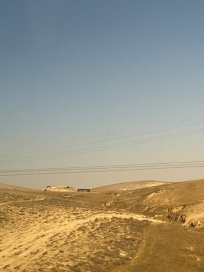 The image depicts a landscape with a clear, pale blue sky and a mountainous, rocky terrain on the right side. Several power lines stretch horizontally across the sky, disappearing into the distance. There are small, dark specks in the sky, possibly birds or debris. On the rocky hillside, there are faint signs of structures or man-made objects, including what appears to be a small building or vehicle. The lighting suggests it is daytime, with the sun casting a warm glow on the rocks. The terrain is arid and appears to be a desert or semi-desert environment. The overall impression is one of a remote, undeveloped area.