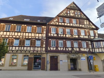 The image shows a street view of the historic town center of Neuenstadt am Kocher, Germany. The focus is on a traditional half-timbered building with a tiled roof. The building has multiple stories with numerous windows, many featuring wooden shutters. The lower facade of the building contains commercial establishments, including an "Apotheke" (pharmacy) with large display windows and a prominent sign. Adjacent to the pharmacy, there is a large wooden door and other entrances to shops or offices. Signs for "Lotto," "Service Point Hoffmann," and a DHL postal box are visible. In the background, a street sign indicates "Metzgerei Lohmann" and "Gasthof Ochsen," suggesting these are nearby establishments. The street in front of the building is paved with cobblestones. The sky is blue with some clouds. The overall scene depicts a well-preserved historical urban setting.