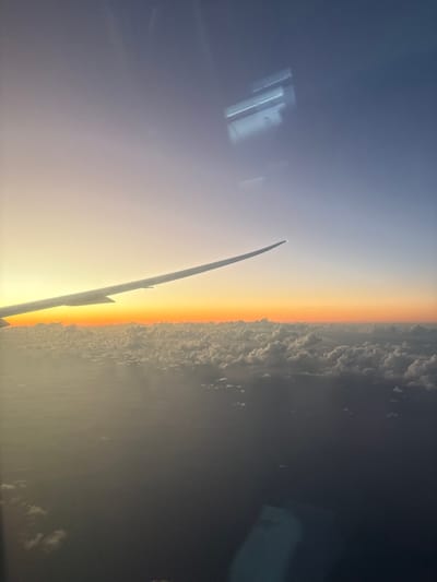 A view from an airplane window captures a vibrant sunrise or sunset. The wing of the plane is visible, silhouetted against the light. Below the wing, a layer of fluffy clouds meets the horizon, which is aglow with orange and yellow hues. Above the horizon, the sky transitions to a darker blue. Reflections of interior lights can be seen in the window.