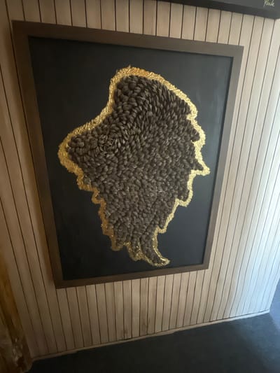 This image shows a framed artwork hanging on a horizontally paneled wall. The artwork itself is abstract, featuring a dark, textured mass outlined with gold leaf against a black background. The textured mass is composed of numerous small, dark, pointed elements, possibly feathers or pinecone scales, arranged in a dense pattern. The gold leaf border is irregular and jagged, highlighting the perimeter of the textured mass.

The wall behind the artwork is made of light-colored, horizontal wooden planks with dark lines separating them, giving it a striped appearance. To the left of the main artwork, a portion of another framed piece is visible, with some handwritten text in cursive on its black surface. To the right of the main artwork, the wall continues, and there's a suggestion of a darker, unadorned wall or corner in the far right of the frame.

The lighting suggests an indoor setting, likely a gallery or a home interior. There are no people visible in the image, and the focus is solely on the artwork and its mounting. The overall impression is one of an art display, with the unique texture and gold accents of the central piece being the main point of visual interest. The setting appears modern and minimalist, allowing the artwork to stand out. There is no indication of the time of day or specific weather conditions. The location context of Portsmouth, United Kingdom, does not provide any specific visual cues within the image itself.