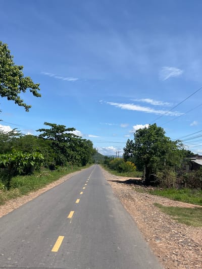 The image shows a paved road with a dashed yellow line marking the center. The road is bordered by green vegetation on both sides, including trees and bushes. In the distance, a mountain range is visible under a clear blue sky with scattered white clouds. The scene appears to be a rural or semi-rural area. There are no people or vehicles clearly visible on the road. The environment suggests a warm climate. The presence of utility poles and wires indicates that this road is in a developed area, likely a village or town. Based on the provided context, this scene is located in the city of Hong Son, Vietnam.