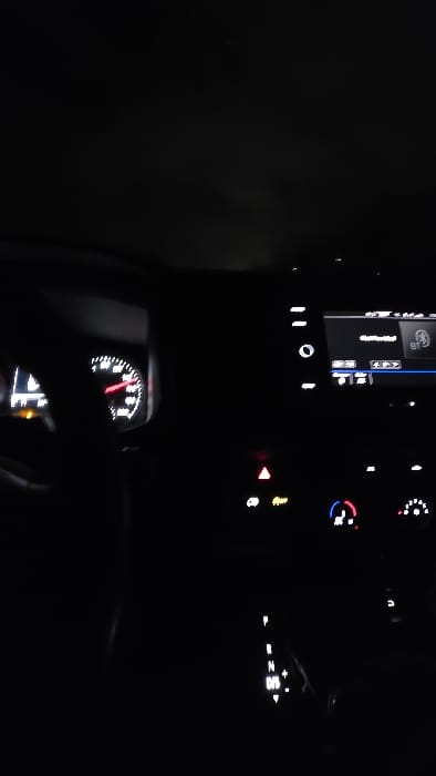 A nocturnal interior view of a vehicle is depicted, showcasing the dashboard and center console within the city of Vorst, Belgium. The scene is predominantly dark, illuminated by the vehicle's internal systems. On the left, a dashboard gauge indicates a reading between 20 and 30 units, with an adjacent illuminated indicator light. The central infotainment screen displays a Bluetooth icon and media information. Below it, illuminated controls include an active red triangular hazard light indicator and climate functions. An automatic gear selector is visible in the lower right, with the 'D/S' position illuminated. An indistinct, dark form, consistent with a human arm or silhouette, is partially visible on the far left. The external environment remains obscured by darkness.