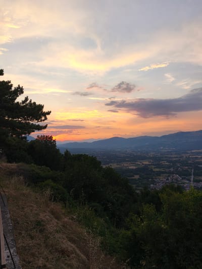 This is a vertical, medium shot of a sunset over a valley with mountains in the background, viewed from an elevated position in Bojano, Italy. The foreground on the left shows a steep, dry grassy embankment with scattered brown stalks and a dark wooden railing or bench along the top edge. Dark green trees and foliage occupy the middle ground, extending into the lower right of the frame. The sky is filled with soft, diffused light from the setting sun, casting hues of orange, yellow, pink, and lavender across scattered clouds. The middle distance reveals a valley floor with agricultural fields, patches of trees, and a small town with red-tiled roofs and a visible road. Distant, hazy blue mountains form the horizon line, and faint wind turbines can be seen on the ridge of the farthest mountain range. The scene is tranquil and natural.
