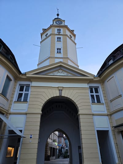 The image shows a low-angle view of the Ansbach city hall tower and its adjoining archway in Ansbach, Germany. The tower is tall and octagonal, painted in cream and yellow with white accents. It features a clock face on one side and several rectangular windows. At the very top, a weather vane is visible. The archway, which leads into a street, is large and curved, supported by substantial pillars. The buildings flanking the archway have curved, tiled roofs and are painted in pale yellow and cream. Several windows with white frames are visible on these buildings. The sky above is a clear, pale blue. The street visible through the archway shows more buildings with shops, awnings, and some outdoor seating. The overall scene suggests an urban setting, likely in a historic European town.