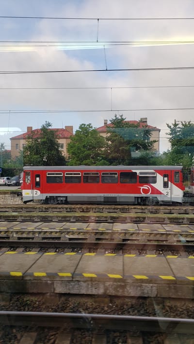 A red and white train car is stopped at a station in Margecany, Slovakia. The train car has a number "2" displayed on both ends. The station platform is visible with yellow and black hazard stripes. In the background, there are buildings with red roofs and green trees. The sky is partly cloudy.