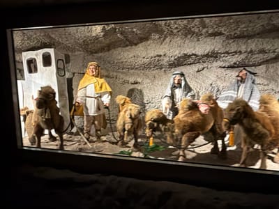 The image depicts a scene in the city of Tolk, Germany, likely at the Tolk-Schau amusement park, which features dioramas and attractions. The scene itself is a diorama of a biblical Nativity scene, rendered in a static, theatrical style.

In the foreground, to the left, is a large, tan-colored camel with a saddle, standing on a sandy ground. Behind it, a figure dressed in a white robe and a yellow head covering, possibly representing Mary, stands near a small, whitewashed structure with two rectangular openings. Further to the right, two more camels, also tan-colored and appearing to be real taxidermy or highly realistic models, are positioned.

The middle ground features two male figures dressed in traditional Middle Eastern attire, including keffiyehs (headscarves). One figure appears to be interacting with the camels, while the other stands slightly behind. All figures are still, suggesting they are mannequins or figures within a display.

The background is a rough, rocky, cave-like wall, creating an austere environment. The lighting appears to be artificial, casting shadows that enhance the dramatic presentation of the scene. The overall impression is of a staged tableau meant to evoke a historical or religious narrative.