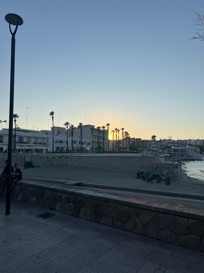 The image shows a coastal scene in Otranto, Italy, at dusk. A pathway paved with stone tiles is in the foreground, bordered by a stone wall. Behind the wall is a sandy beach. Several palm trees are scattered across the scene, some silhouetted against the sky. Buildings line the waterfront, appearing to be hotels or businesses with balconies and awnings. The sun is setting, casting a warm glow on the horizon and creating a light haze. On the beach, there are a few people, including two individuals sitting on the wall in the foreground, and a small group near a green tent in the middle ground. The sea is visible to the right, with gentle waves lapping at the shore. A lamppost stands prominently on the left side of the image, partially obscuring the view of the buildings.