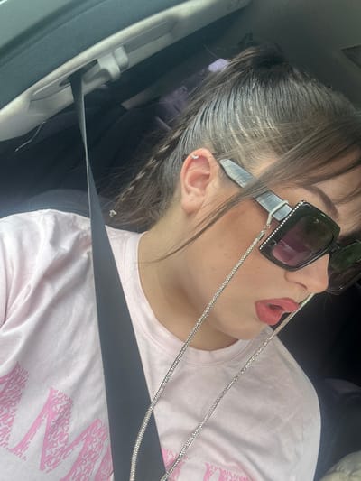 A young woman, likely in her late teens or early twenties, is captured from the chest up in a close-up, side-profile shot. She is seated in the passenger seat of a vehicle, indicated by the visible seatbelt and the interior of the car. The woman has her hair pulled back in a high ponytail, with a braid visible. She is wearing large, square-shaped sunglasses adorned with rhinestones, and a diamond-encrusted chain is attached to them, draped across her chest. Her lips are painted a bold red. She is wearing a light pink t-shirt with a large, stylized graphic in a bubble-like pink font that appears to be an abstract design. A black seatbelt runs diagonally across her torso. The lighting suggests it is daytime, and the image was taken indoors, likely within a car. The location is identified as Dolna Banya, Bulgaria, although this context is not visually discernible from the image itself.