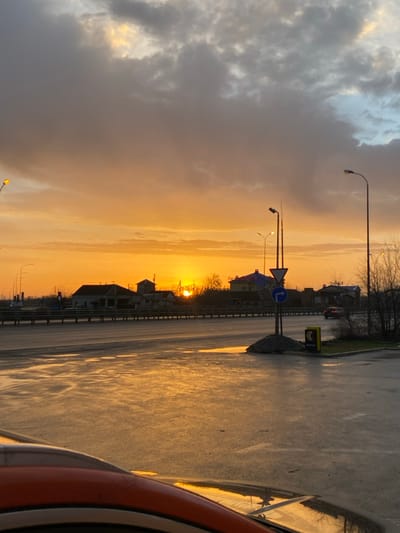 A street scene at sunset, taken from inside a vehicle. A wet, reflective road stretches into the distance, reflecting the warm hues of the setting sun. Houses line the street, silhouetted against the orange and yellow sky. Streetlights and road signs are visible along the road. A storm cloud hangs to the left, contrasting with the bright sky and adding dramatic depth to the scene. Part of the vehicle's interior is visible on the right.