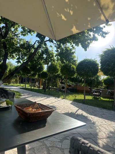 The image depicts an outdoor dining area in Ashtarak, Armenia, likely at a restaurant or cafe. The foreground shows a dark, possibly metal table with a wicker basket containing sliced bread. To the left of the basket, a plate holds a green bell pepper and some white cheese. In the background, a stone pathway winds through a lush garden with manicured trees and a large, leafy tree canopy overhead, partially obscured by a cream-colored umbrella. Sunlight streams through the leaves, casting dappled shadows on the pathway. Several outdoor seating areas with wicker furniture are visible in the middle ground, along with a low wooden fence and decorative elements like a large clay amphora. The overall atmosphere is tranquil and serene, suggesting a pleasant daytime setting. No people are visible in the image.