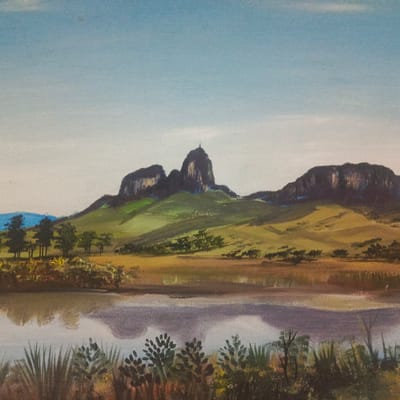 The image is a painting depicting the Guatopo National Park, located near the city of Los Teques, Venezuela. In the foreground, a body of water reflects the sky and the surrounding vegetation. The middle ground features a grassy plain dotted with small trees and bushes, leading up to a series of rolling hills covered in green and yellow vegetation. Dominating the background are large, dark rock formations, with a prominent peak topped by a cross. The sky is a gradient of light blue, with hints of white clouds or haze. The painting style is realistic, with visible brushstrokes and a somewhat muted color palette. There are no people or animals depicted in the scene.