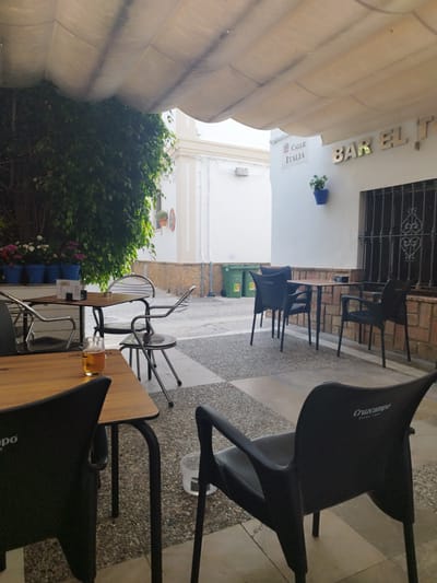 The image depicts an outdoor seating area of a bar, possibly located in Rota, Spain, as indicated by the "CALLE ITALIA" sign and the "BAR EL" signage on the building. The scene is viewed from a low angle, looking towards a white building with a wrought-iron window. A white awning stretches over the seating area, providing shade.

In the foreground, a wooden table with black chairs is visible. One of the chairs is a black plastic chair with the "Cruzcampo" logo embossed on its back. A partially filled glass of beer sits on the wooden table.

Further back, more tables and chairs are arranged on a patterned tiled floor. To the left, a dense green hedge or plant covers a portion of the wall. In front of the hedge, several blue pots containing flowers are lined up. Two green recycling bins are visible in the background. The building itself has a white facade with a brick accent at the base. The overall atmosphere suggests a casual and relaxed outdoor dining or drinking establishment.