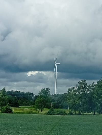 This is a photograph of a landscape in Kalniškiai, Lithuania, under a cloudy sky. In the center of the frame, a tall white wind turbine stands with its blades extended, appearing to be in motion against the dark, dramatic clouds. The turbine is situated behind a line of lush green trees that form a dense forest in the middle ground. In front of the trees, there is a green field, possibly a meadow or crop field, with some white posts visible. The foreground is dominated by another green field, blurring into the distance. The overall impression is one of a rural, natural setting with renewable energy infrastructure.