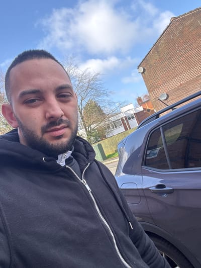 The image displays a male individual in the foreground, engaged in what appears to be a selfie, outdoors on a residential street in Preston, United Kingdom. The man possesses short dark hair and a beard, and is wearing a black zip-up hooded sweatshirt, revealing a white garment and a silver-colored chain or pendant beneath its opening. To the subject's right, a dark grey SUV with roof rails and tinted rear windows is partially visible, parked on the street. The background features a brick building with two satellite dishes affixed to its exterior wall. Further into the background, bare deciduous trees and additional residential structures are discernible, including one with white walls and another exhibiting patterned brickwork. A light-colored wooden fence demarcates a property line in the mid-ground. The scene is illuminated by natural daylight under a blue sky with scattered white clouds.