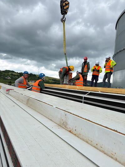 The image shows a group of construction workers wearing hard hats and high-visibility vests on a rooftop, with a cloudy sky overhead and a rural landscape in the background. A crane hook and yellow strap are visible, indicating construction activity, likely lifting or positioning materials. The workers appear to be engaged in their tasks, with some looking at their phones or coordinating. The rooftop is covered with large, white, panel-like structures, suggesting the construction of a building or industrial facility. Based on contextual information, this scene is located in Jodłownik, Poland.