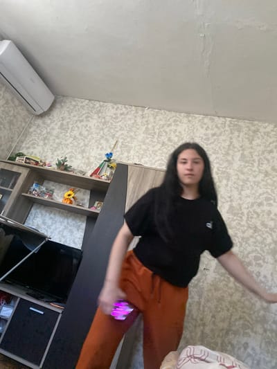 A young person with long, dark hair stands in a room, holding a spinning, illuminated toy. They are wearing a black t-shirt and orange pants. The room has light-colored walls with a textured wallpaper pattern. To the left, a shelf unit contains decorative items, including a small pumpkin, flowers, and framed pictures. An air conditioning unit is mounted high on the wall above the shelves. A television and a dark cabinet are visible in the lower left portion of the image. The overall environment suggests a domestic setting within a residential building in Ovcha Mogila, Bulgaria.