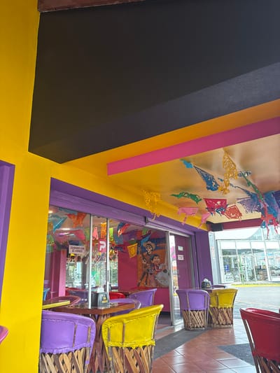 The image shows the interior of a brightly colored restaurant in Pozos, Costa Rica (9.951, -84.194). The walls are painted yellow and purple, and the ceiling is black, pink, and yellow with brightly colored paper decorations hanging from it. There are several tables and chairs set up in the restaurant. The tables are made of wood and the chairs are woven from natural materials. There is a sign in Spanish that says "cuidado con el vidrio." There are several large windows that look out onto a street scene. There is a picture of a cartoon character on the wall, possibly from a popular Mexican animated film. The restaurant appears to be open for business. There are no customers visible in the image. The image is taken from the viewpoint of someone standing inside the restaurant.