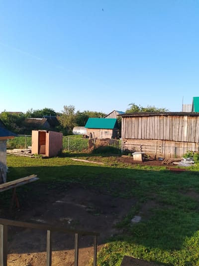 The image shows a backyard scene in Kranokholmskiy, Russia, on a clear day with a bright blue sky. In the foreground, there is a dark wooden railing and some wooden planks resting on a support. The middle ground features a grassy yard with some muddy patches, a small wooden shed under construction, and a larger wooden structure with a corrugated metal roof. Behind these structures, a chain-link fence separates the yard from a garden area with rows of plants and a greenhouse. Several other buildings with various roof types, including green and grey corrugated metal, are visible in the background, surrounded by trees and vegetation. A single bird is captured in flight in the upper left portion of the sky. The scene appears to be a residential or dacha property, suggesting a rural or semi-rural setting.