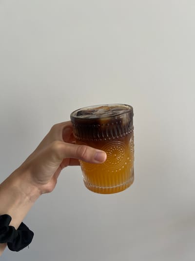 A hand holds a glass tumbler at an angle against a plain white background. The tumbler contains a two-layered beverage with dark brown liquid at one end and a lighter orange liquid filling the majority of the glass. Some ice cubes are visible inside the tumbler. The tumbler is made of glass, has a rounded, cylinder shape, and features an embossed design around its circumference. The hand holding the glass has fair skin. A black scrunchie is visible around the wrist of the hand. The background is a uniform, light grey color. The location is impossible to determine based on the image content.