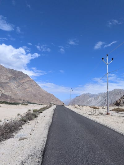 The image depicts a paved road winding through a dry, mountainous landscape under a bright blue sky with scattered white clouds. On the left, a large, layered mountain with rough, arid terrain dominates the background. The roadside is sandy and sparsely vegetated with dry bushes and scattered rocks. On the right side of the road, a telephone pole with multiple wires stretches across the frame, leading towards distant, rugged mountains. Further down the road, another telephone pole is visible. A single motorcyclist is seen riding away from the viewer on the road. The environment appears to be a remote, high-altitude region in India, potentially Sasoma, known for its challenging terrain and stark beauty.
