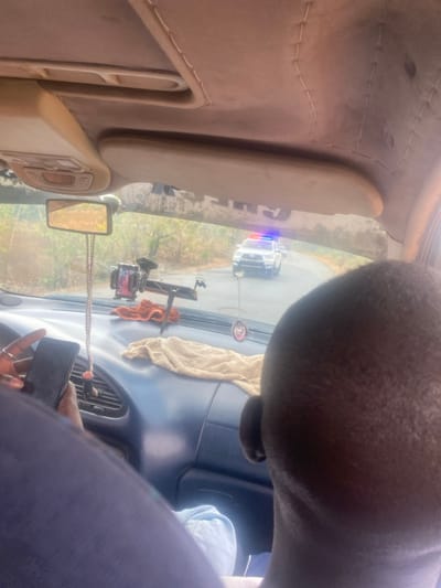 The image depicts the interior of a vehicle, viewed from a rear perspective, looking forward through the windshield. In the immediate foreground on the right, the back of an individual's head with dark skin and closely cropped hair is visible. On the left, a hand with dark skin, adorned with a ring, holds a black smartphone. The vehicle's dark blue dashboard features a beige towel, a black phone mount, and a decorative item suspended from the rearview mirror. Through the windshield, a white pickup truck, equipped with active blue and red emergency lights on its roof, is visible on an asphalt road ahead. The roadside environment consists of sparse, dry vegetation. This scene is located in Nandu, Nigeria.