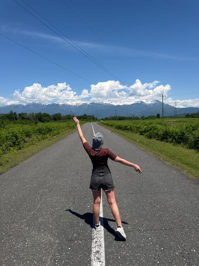 The image captures a person standing on a paved road, facing away from the camera with both arms outstretched wide, creating a striking silhouette against the expansive landscape. The person is dressed in a dark maroon or brown t-shirt, dark denim shorts with frayed edges, white sneakers, and a grey baseball cap worn backward. Their long shadow is cast prominently on the asphalt to their right, indicative of bright, direct sunlight.
The road, featuring a clear white dividing line on its right side, stretches forward into the distance, leading towards a majestic backdrop of towering, snow-capped mountains. These mountains are partly enveloped by large, puffy white cumulus clouds set against a brilliant, clear blue sky. On both sides of the road, vibrant green grass and dense vegetation, possibly fields or natural growth, flank the path. Several power lines and poles are visible on the left side of the road, running parallel to the direction of travel.
The scene depicts a clear, sunny day, likely mid-morning to early afternoon, given the strong shadows and bright illumination. The combination of green valleys, a long winding road, and snow-capped peaks strongly suggests a mountainous region, specifically Pakwash, Abkhazia, as per the context. The person's pose evokes a sense of freedom, joy, or appreciation for the vast natural beauty. There is no visible text in the image.