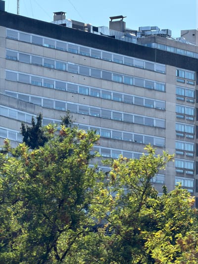 The image shows the facade of a multi-story building in Bron, France. The building appears to be an older, institutional structure, possibly a hospital or office building, constructed from concrete panels. Numerous windows, covered with horizontal blinds, are arranged in rows across the facade. The rooftop features visible HVAC units and ventilation systems, as well as chimneys. In the foreground, lush green trees with some autumnal foliage obscure parts of the building, suggesting the photo was taken from a street or park adjacent to the structure. The sky is clear and blue. There are no people visible in the image.