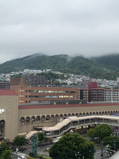 An elevated, wide-angle view captures an urban landscape in the city of Takarazuka, Japan, under an overcast sky.

In the foreground, a multi-lane roadway is visible, with a white passenger vehicle positioned near a pedestrian crosswalk on the left. A dark, multi-faceted sculptural object stands adjacent to the crosswalk. Above the street, an extensive network of beige-colored elevated pedestrian walkways, featuring translucent arched roofs and staircases, connects various buildings. Several individuals are discernible on these walkways and at street level.

The midground is dominated by a large, multi-story, light-tan building with a terracotta-colored roof. This structure, part of a station complex, displays Japanese characters (駅 - "Eki," meaning station) on its facade and features a long series of arched windows on a lower level. Behind and to the right of this building, a brown brick building with the name "SOLIO 2" (ソリオ2) visible on its roofline presents multiple rectangular windows, some of which are illuminated. To the right, a multi-story residential or commercial building with a red roof section exhibits numerous balconies and windows arranged in a grid pattern.

The background consists of densely forested mountains, their peaks and upper slopes partially obscured by low-hanging mist or clouds. Numerous residential houses and smaller buildings are visible across the lower and middle slopes of these mountains. Several thin, vertical antenna or communication towers are positioned on the mountain peaks. The sky above is uniformly gray, indicating full cloud cover.