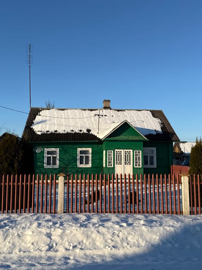 A green, two-story house with white-framed windows stands beside a brown, slatted fence. The roof is partially covered in snow. An antenna stretches toward the clear blue sky. The ground is blanketed in snow, with shadows cast by the sun. A small, green bush sits near the house's base, adding a touch of foliage to the winter scene. A street sign can be seen on the house's green facade.