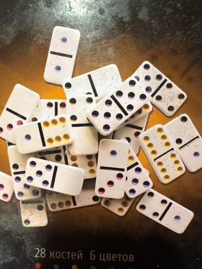 A close-up, top-down shot displays a jumbled collection of dominoes scattered on a dark, textured surface. The dominoes are predominantly white with black dividing lines and colorful pips, including purple, yellow, pink, and green. Some dominoes show signs of wear. In the lower portion of the image, text is visible on the dark surface, reading "28 костей 6 цветов" (28 bones 6 colors) along with colored dots. The scene appears to be an indoor setting, but there are no clear indicators of its specific location within Tsandrypsh, Abkhazia.