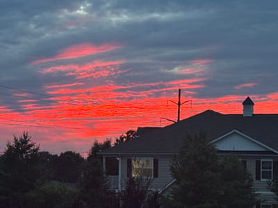 A sunset with vibrant red and orange clouds is visible in the sky over Allen Township, United States of America. The clouds are streaked and horizontal, with darker gray clouds above and below them. In the foreground, the dark silhouette of a house with a dark roof is visible. Trees are also visible in the foreground, obscuring parts of the house. Power lines are stretched across the sky.