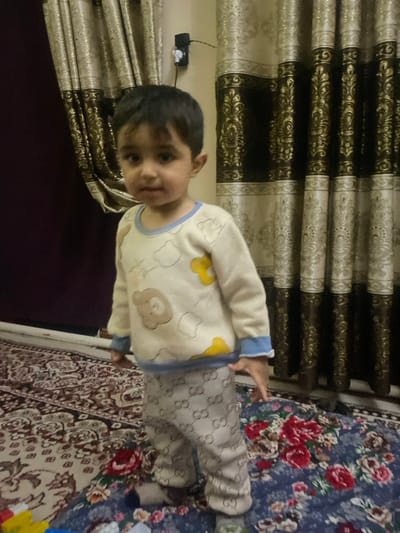 A young child, approximately a toddler, stands facing forward in an indoor residential setting in Galaosiyo, Uzbekistan. The child has dark hair and dark eyes, looking directly at the viewer with a neutral expression. They are wearing a light-colored long-sleeved top with blue trim at the collar and cuffs, featuring multiple cartoon bear graphics and other light-colored shapes. Below, the child wears light-colored pants patterned with repeating "GG" logos. The background consists of pleated curtains; the right side features dark brown and light gold ornate patterned curtains, while the left side shows a darker, possibly deep purple or burgundy curtain. A light-colored wall section is visible between the curtains on the upper left, featuring a black electrical outlet with a white plug adapter. The floor is covered by two layered rugs: a prominent dark blue rug with large red and pink floral patterns is in the foreground and to the right, overlapping a lighter-colored rug with an intricate traditional pattern in shades of cream, maroon, and brown visible on the left and center. The child's hands are down by their sides, and no interaction with other individuals or specific objects is observed.